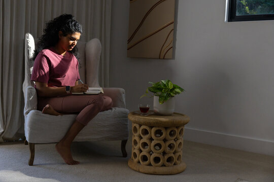 Woman sitting chair at home writing notebook, pen, wearing mauve outfit, smartwatch, side table cup