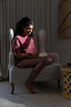 Woman sitting curled in wingback armchair at home by curtained window writing in notebook with pen