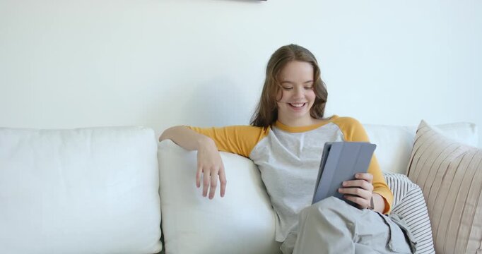 Female sitting on white couch in raglan shirt, using folio tablet, reacting to content, copy space