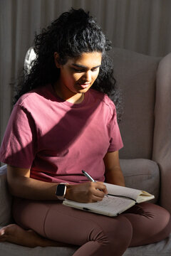 Female sitting cross-legged on armchair at home, writing in open notebook with pen and smartwatch