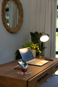 Wooden desk is hosting open laptop, glowing brass lamp, potted plant and teacup near window