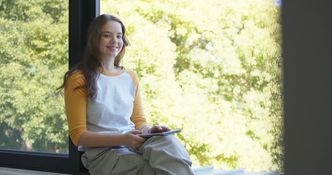 Female sitting on windowsill holding tablet glancing at camera smiling shifting legs, copy space