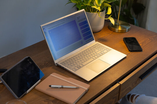 Open laptop resting on warm wooden desk with tablet, notebook, phone, plant, lamp, denim-clad leg