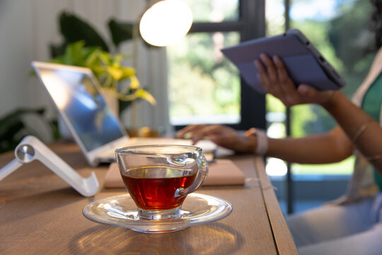 Indian woman multitasking at home office desk, using tablet and sipping dark tea from glass teacup