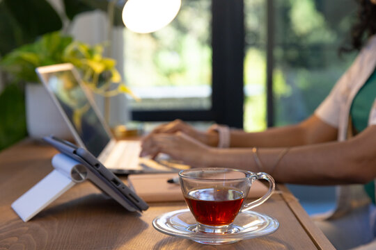 Clear glass teacup is resting on wooden desk, steaming amber tea near tablet and laptop