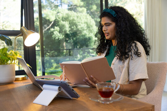 Woman holding open notebook, speaking to laptop on wooden desk by window with tea