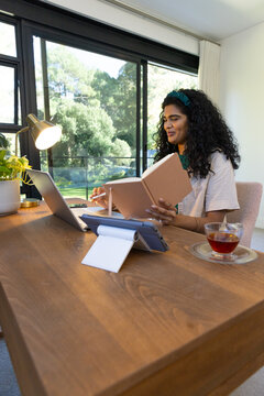 Indian woman holding notebook and pen at home office desk, using laptop with tea, copy space