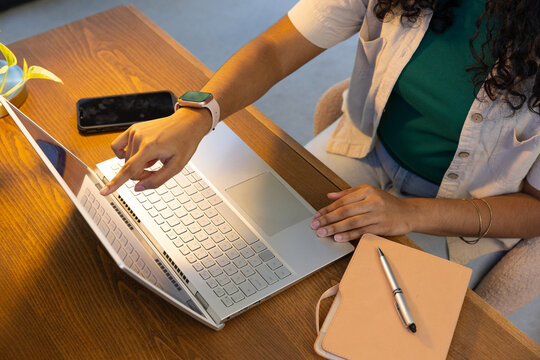 Indian woman pointing at silver laptop while typing at wooden desk with phone, wearing green top