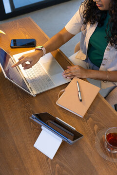 Woman in green top pointing at silver laptop on wooden table with notebook, smartphone, copy space