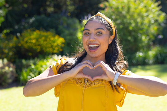Woman smiling and making heart with hands standing on sunny lawn wearing yellow top headband watch