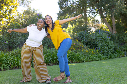 Two adult women posing in garden, linking arms and smiling with yellow headband and striped pants