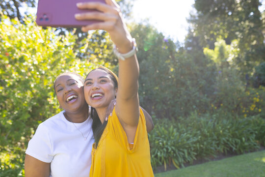 Diverse female friends leaning together, taking selfie in sunlit garden with pink smartphone case