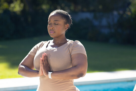 African American woman praying at backyard pool, wearing beige T-shirt and white smartwatch