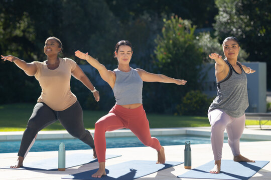 Diverse female friends in sportswear practicing standing pose on pool deck with mats, water bottles