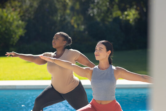 Diverse female friends practicing standing yoga in backyard beside swimming pool wearing leggings