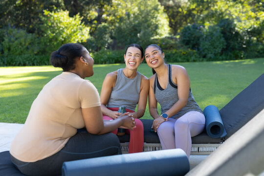 Diverse female friends laughing and chatting in sportswear on patio with foam roller and yoga mat