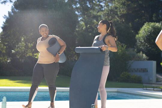 Diverse female friends holding yoga mats and water bottle preparing for outdoor class poolside