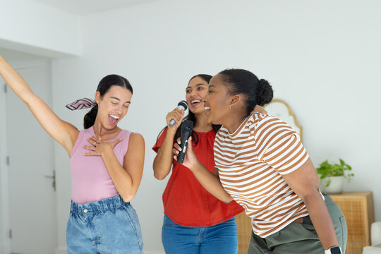 Diverse female friends singing in living room, wearing casual tops with microphones by wood cabinet