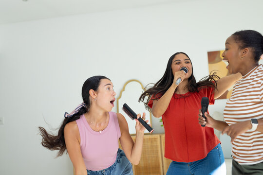 Diverse female friends singing and dancing while holding microphones and hairbrush in living room