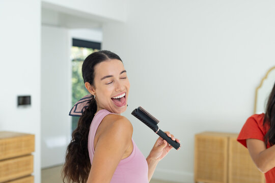 Female friends singing in bedroom, one wearing pink tank top using heated round hair brush