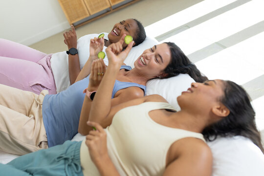 Diverse female friends reclining on bed at home, holding cucumber slices and wearing loungewear