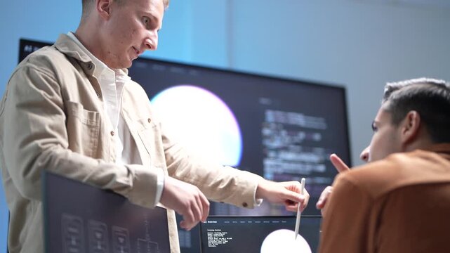 A senior technology architect pointing out system optimization parameters and database structure models on a monitor to a software developer.