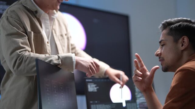 A senior technology architect pointing out system optimization parameters and database structure models on a monitor to a software developer.