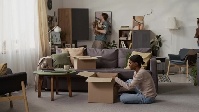 Caucasian young man taking piles of clothing items out of closet and putting them on couch while Black girl labelling cardboard box in living room