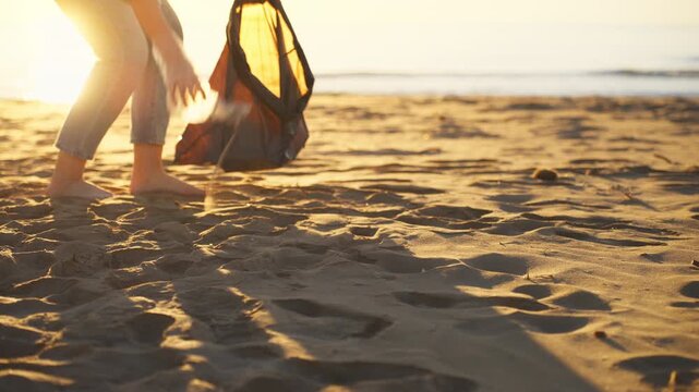 Young volunteer woman walking barefoot on sandy beach during sunset, picking up plastic bottle from sand and putting it in garbage bag for recycling to clean up environment