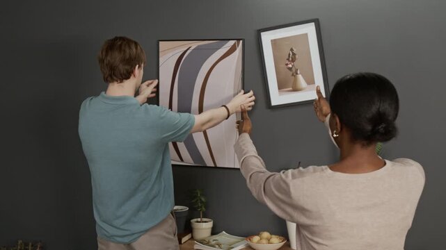 Caucasian young man adjusting alignment of picture frames on grey wall, Black girl watching and guiding him, then they hugging pleased with result