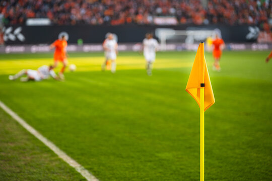A close-up of a bright yellow corner flag on a grass pitch with a football match and stadium crowd blurred in the background.
