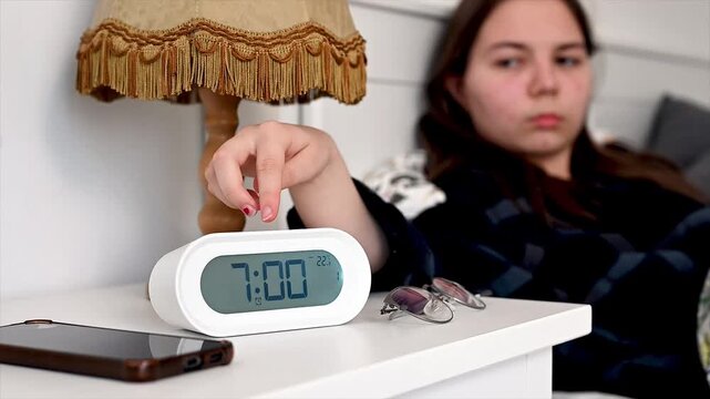 Sleepy teenager reaching for ringing alarm clock on bedside table in morning light, groggy awakening moment and beginning of daily school preparation routine showing relatable adolescent struggle with