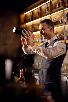 Happy bartender using cocktail shaker while making a drink at bar counter.