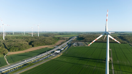 Fototapeta premium Aerial view of a large wind turbine with red striped blades standing next to a busy highway with traffic crossing green agricultural fields under a clear blue sky.