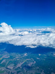 Obraz premium Aerial landscape view from airplane above dramatic white clouds and agricultural fields under bright blue sky. Scenic flight perspective showing countryside patterns and towering cloud formations from