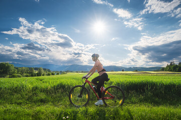 Fototapeta premium Female cyclist riding mountain bike through vibrant green meadow in Slovenia in summer. Woman cycling in scenic countryside landscape under bright sunny sky with lens flare. Active lifestyle. Sport