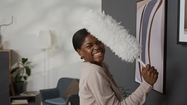 Chest-up portrait of delighted African American gen Z woman cleaning framed artworks with feather duster and posing for camera