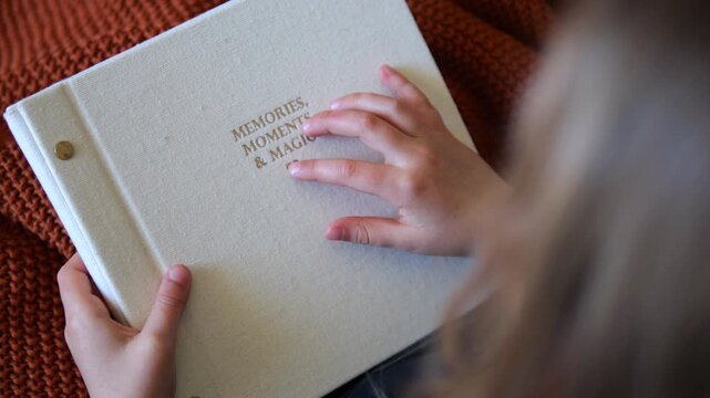 Young girl's hands gently touching the embossed gold lettering on a white fabric photo album, cherishing the magical moments and memories held within the scrapbook in a cozy, warm setting