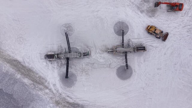 Top-down view of two mobile drilling rigs with dust halos and an orange excavator with yellow bulldozer working on white gypsum mineral surface in open pit quarry