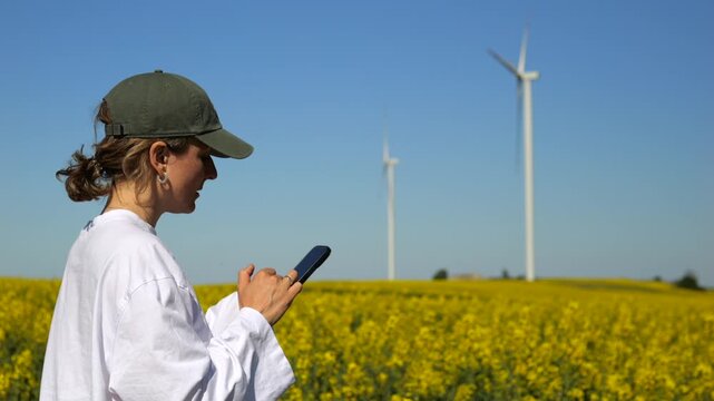 Renewable energy engineer using her smartphone to check wind turbine performance while standing in a yellow rapeseed field, representing sustainable technology and clean power generation