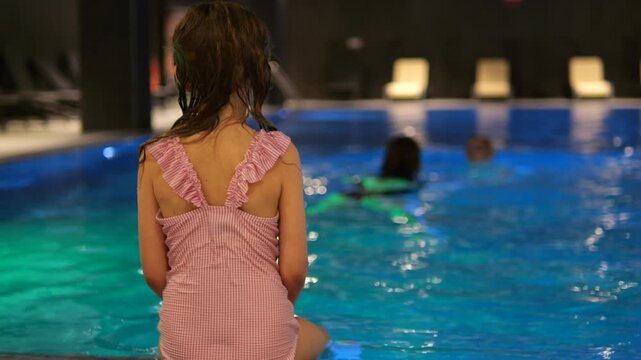 Young girl in a pink dress stands near an illuminated indoor pool, watching swimmers enjoy the water under warm lights as reflections ripple across the calm blue surface