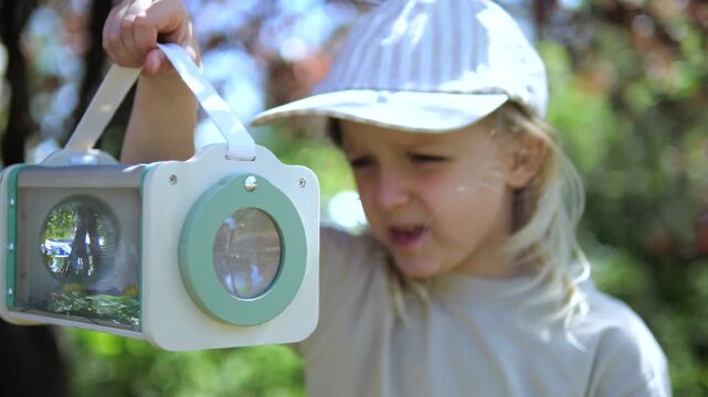 Young female child with a sense of wonder intently observing a captured insect inside a special container with a magnifying lens, learning about entomology and wildlife in a lush green garden