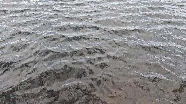 Grey natural water surface filmed at low wide angle in slow motion shows steady small ripples continuously moving across frame under overcast diffused daylight outdoors