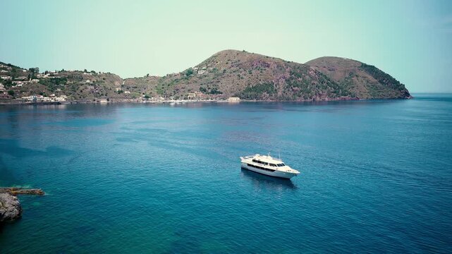 Lipari/Vulcano bay &mdash; white motor yacht in turquoise Aeolian turquoise Mediterranean waters off the coast of Sicily, Italy, with scenic hillside villages and colorful flowers in the foreground.