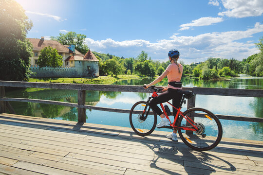 Female cyclist with mountain bike standing on wooden bridge in Slovenia. Woman looking at historic Otocec Castle on Krka river under blue sky. Summer cycling trip, travel destination. Girl on bike
