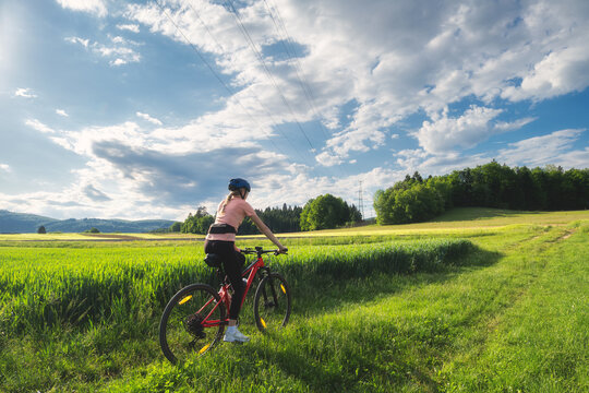 Female cyclist riding mountain bike on gravel path through green fields in Slovenia in summer. Woman cycling away into scenic countryside landscape at sunset. Active lifestyle. Girl on bike. Sport