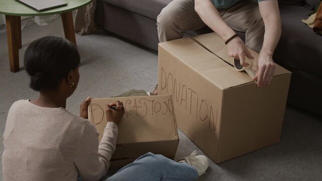 Black girl drawing outline around letters of word donation on cardboard box while unrecognisable Caucasian man sealing another one