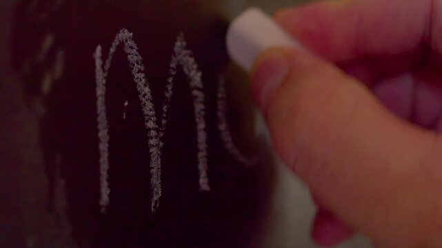 Close up of hand writing illegible unreadable word on chalkboard.A macro shot with shallow depth of field showing a hand using white chalk to write a messy, unknown, or mysterious word on a blackboard