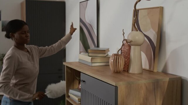 African American young woman cleaning framed art print and wooden cabinet with white feather duster while doing housework