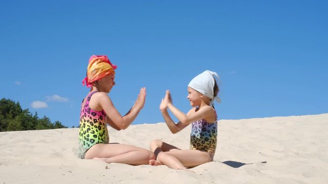 Cute playful girls in swimsuits are playing palm games with each other, sitting on the hot sand of the beach on the coast
